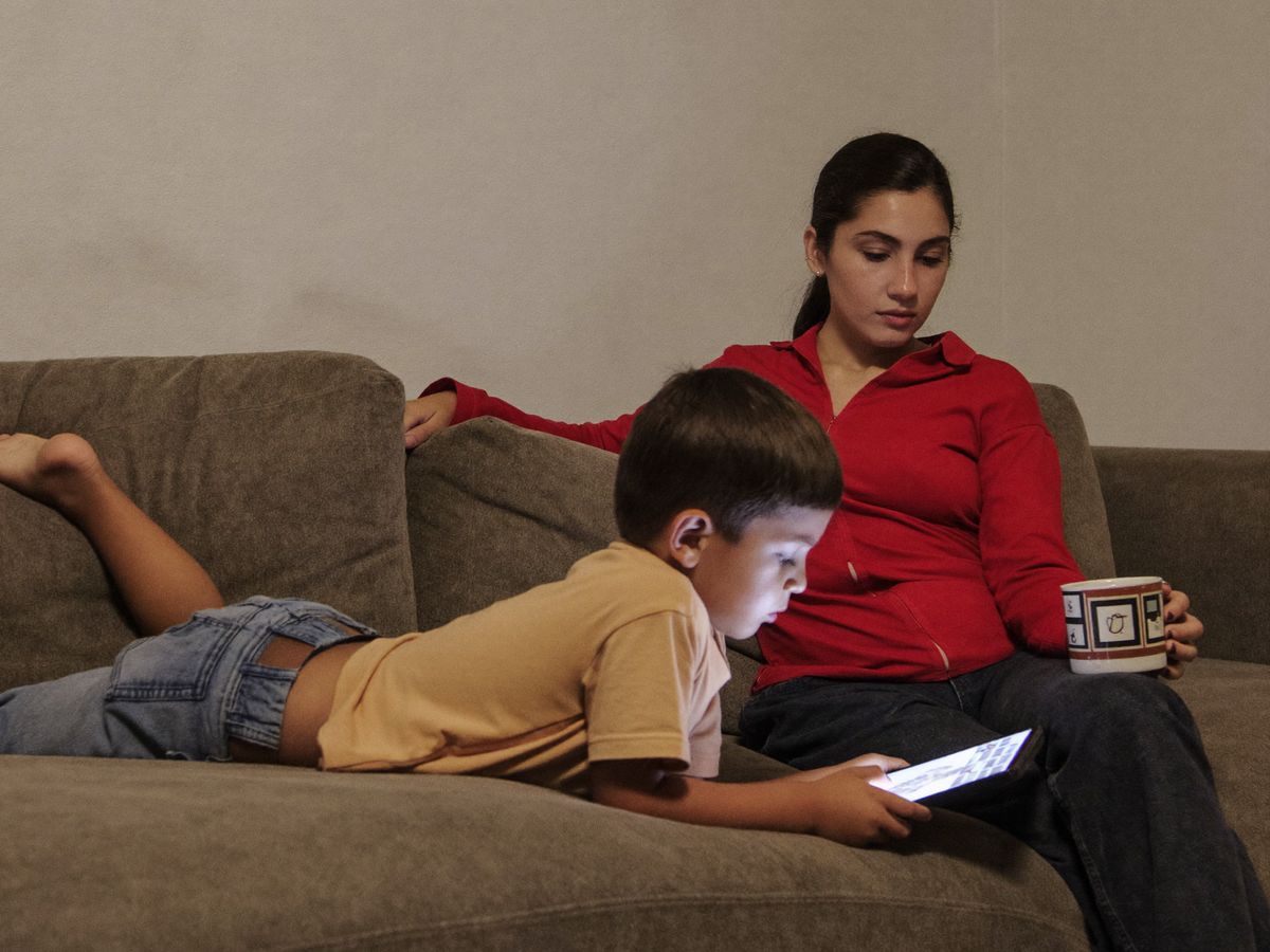 Menino deitado no sofá olhando para o tablet enquanto a mãe, cansada, olha para ele